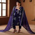 Woman wearing a purple embroidered outfit with a sheer dupatta in a warm-toned room.