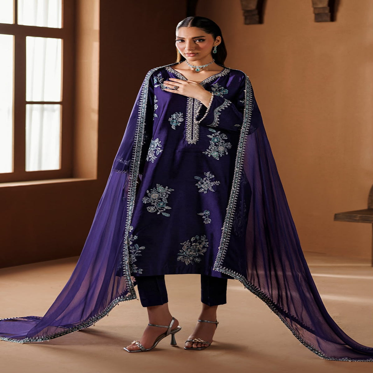 Woman wearing a purple embroidered outfit with a sheer dupatta in a warm-toned room.