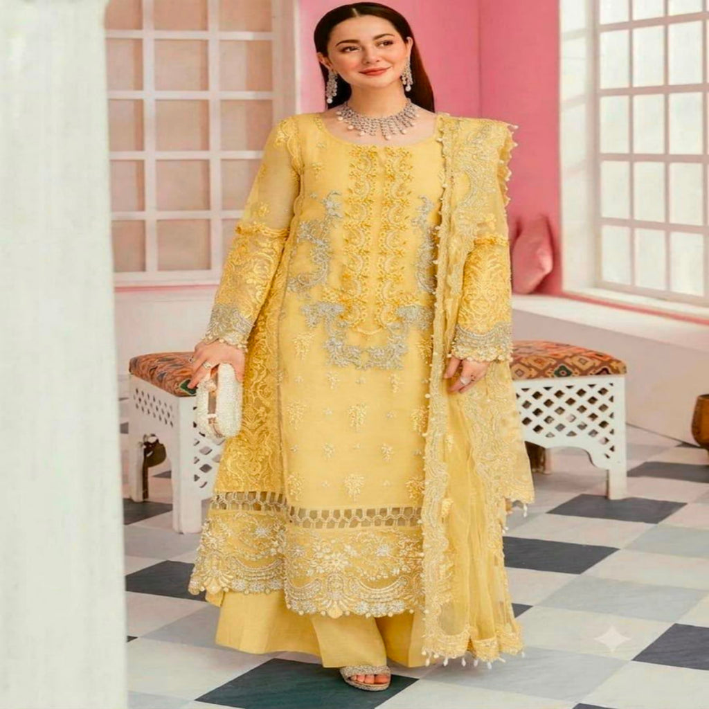 A woman models a traditional three-piece yellow ethnic suit with intricate silver embroidery, featuring a long kurta, matching pants, and a draped dupatta, set against a pink and white interior background.