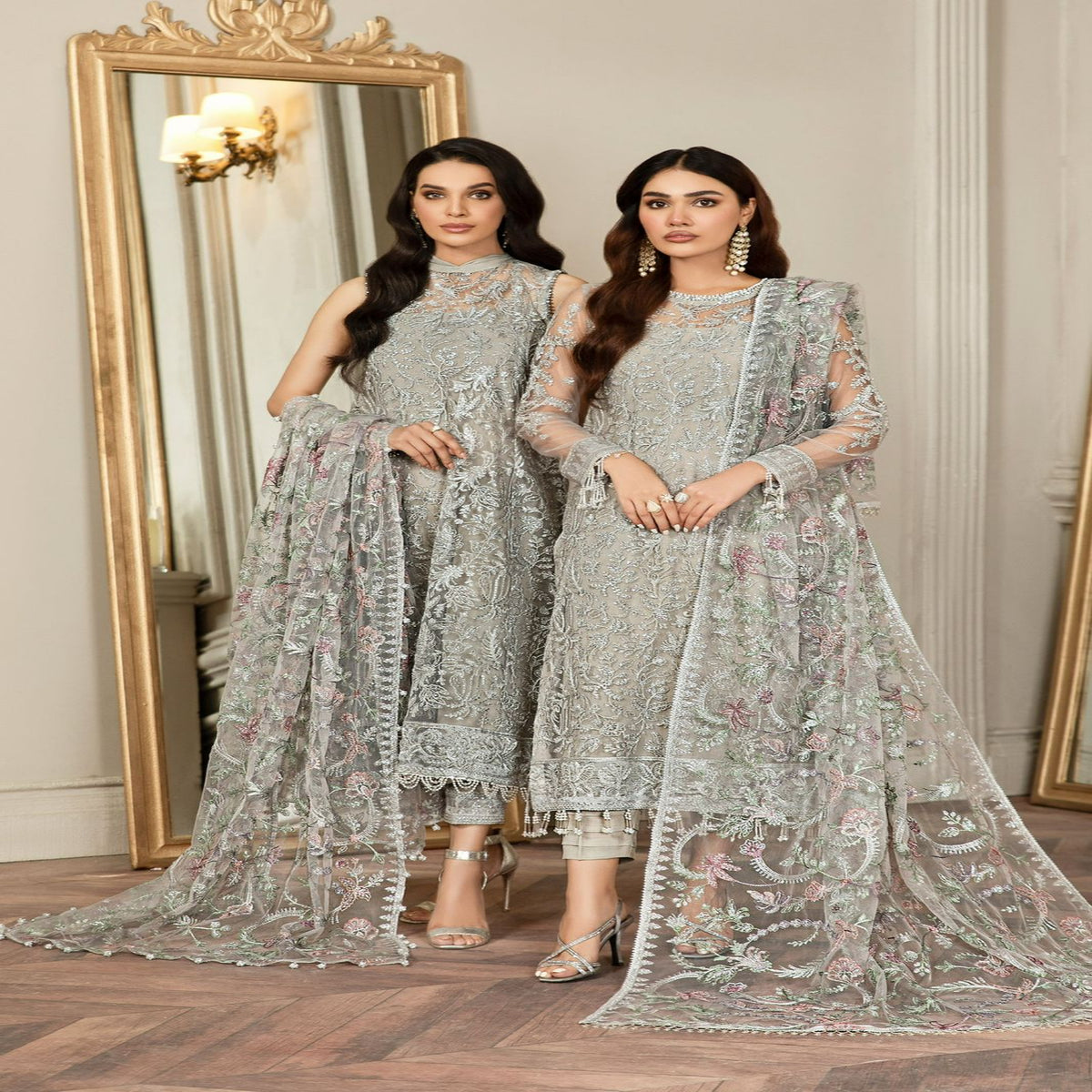 Two women in silver embroidered traditional outfits standing in front of a mirror.