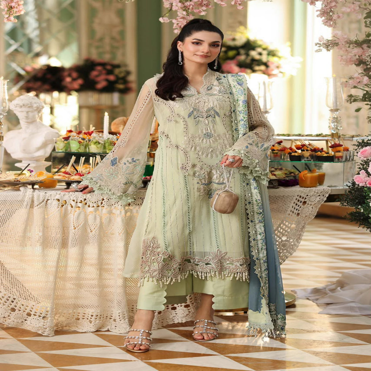 Woman in a light green embroidered outfit standing in an elegant indoor setting with floral decorations.