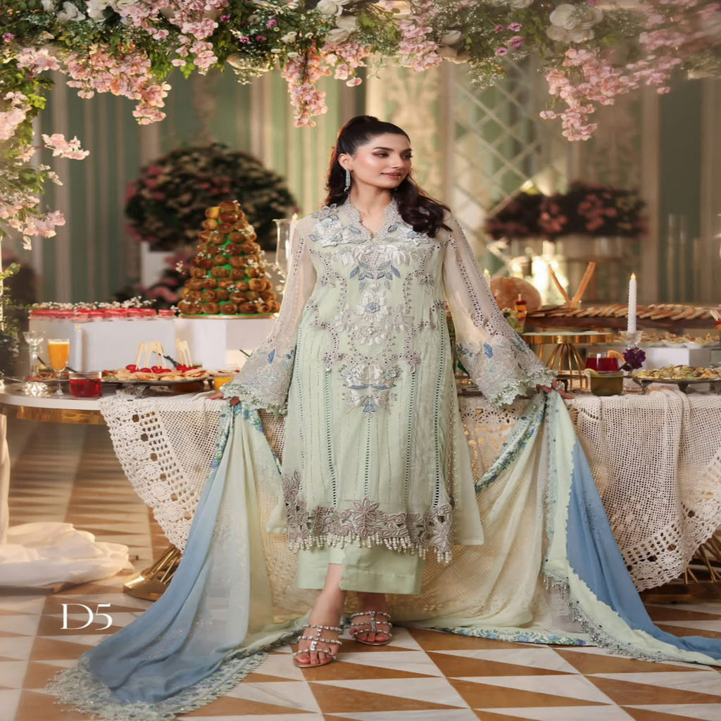 A woman models a traditional South Asian salwar kameez suit in a decorated indoor setting. The outfit is a light sage green with intricate silver embroidery and features a matching light blue dupatta.