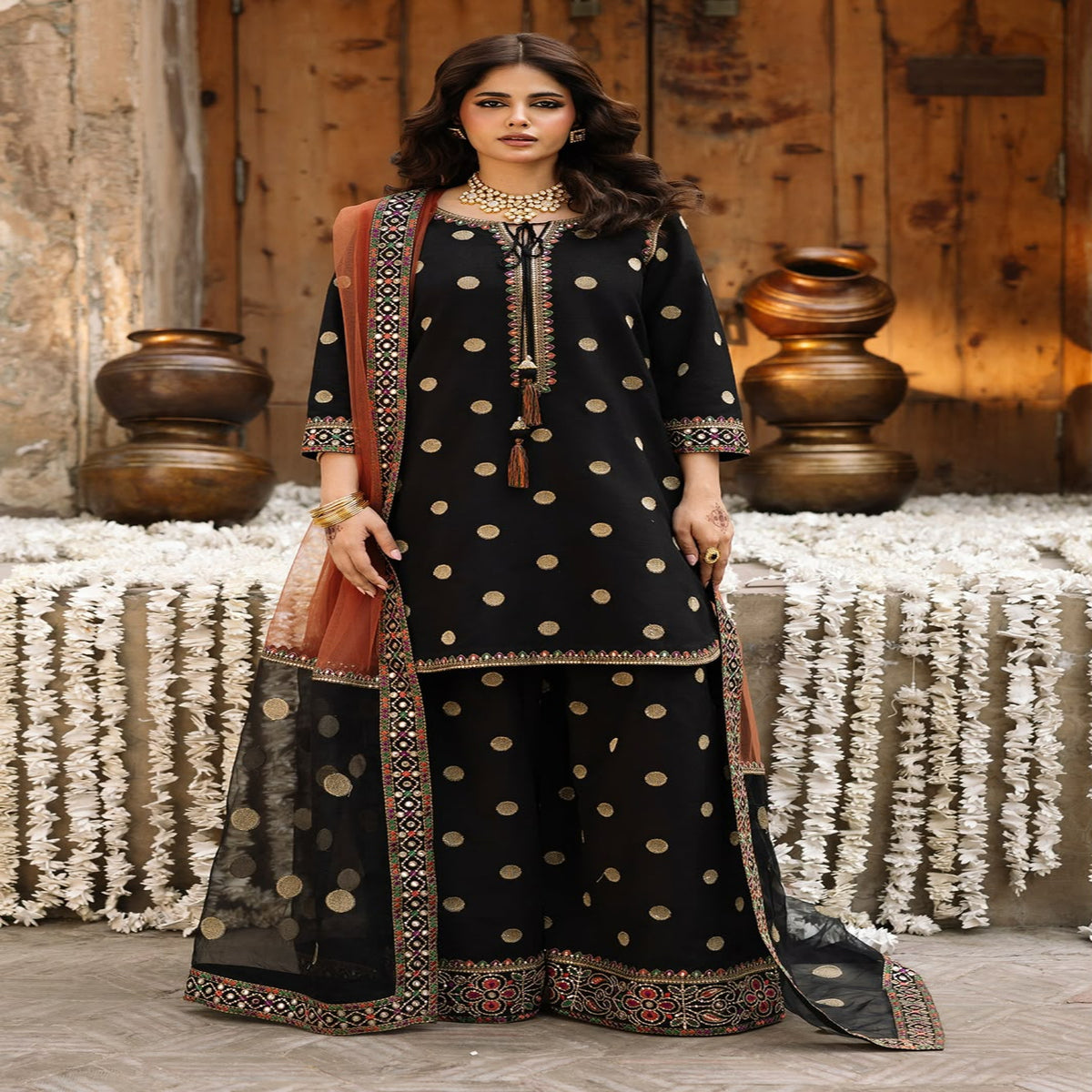 Woman in traditional black and gold outfit with decorative elements in a rustic setting.
