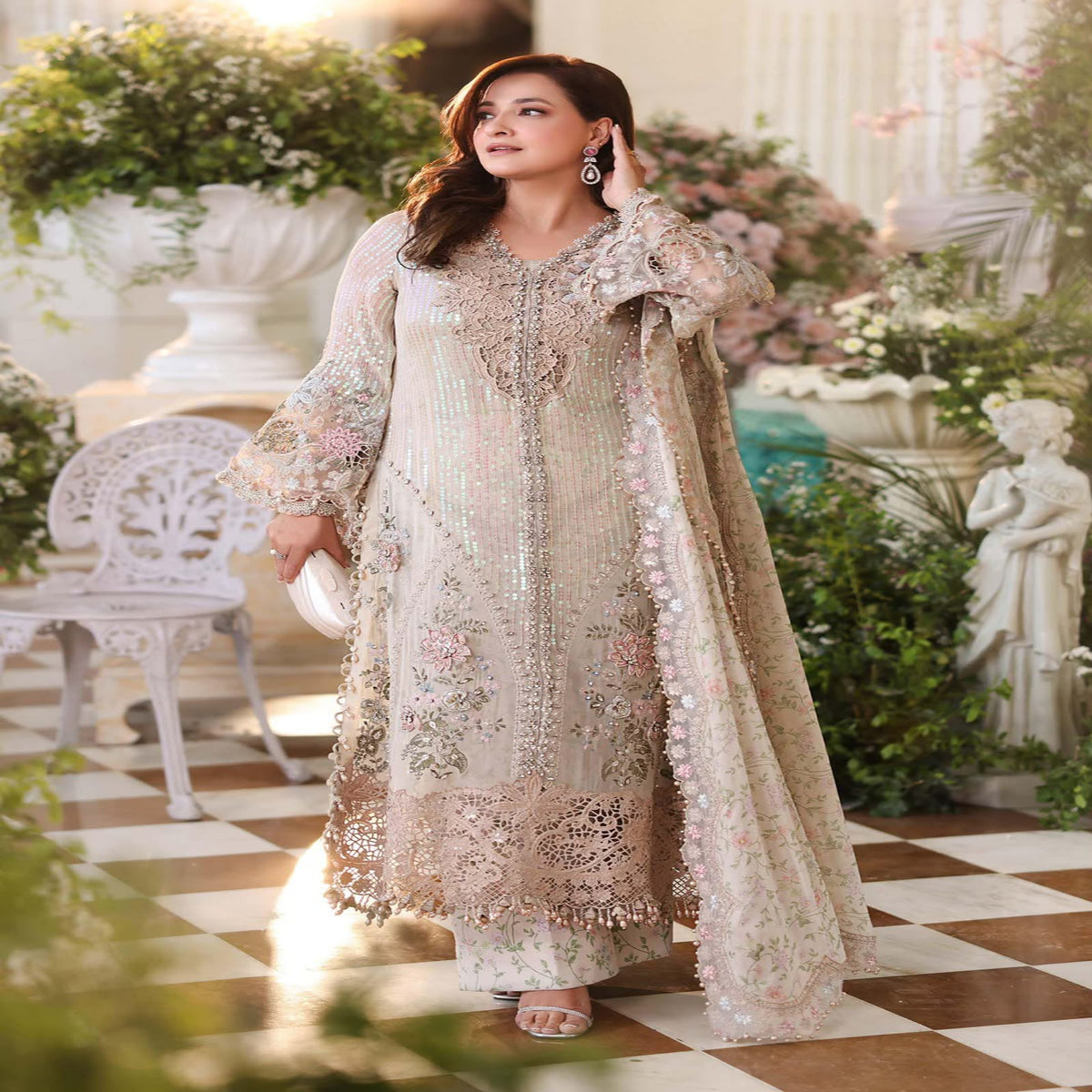Woman in an embroidered outfit standing in a decorative indoor setting with flowers and furniture.