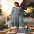 Woman in a light blue embroidered traditional outfit standing in a decorated room with flowers and furniture.
