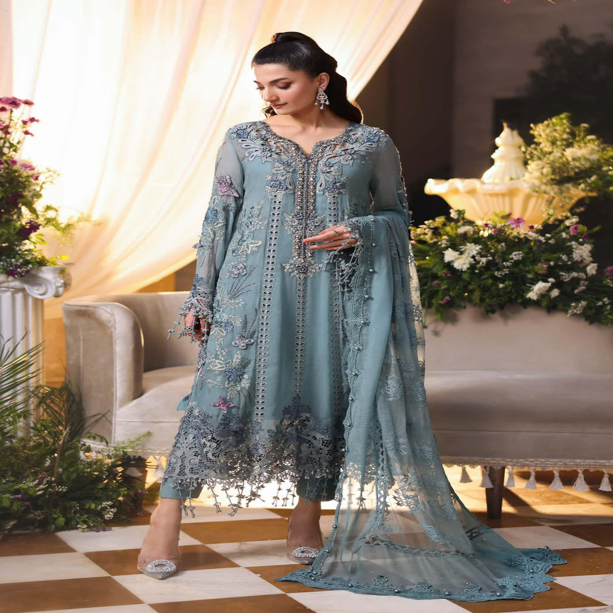 Woman in a light blue embroidered traditional outfit standing in a decorated room with flowers and furniture.