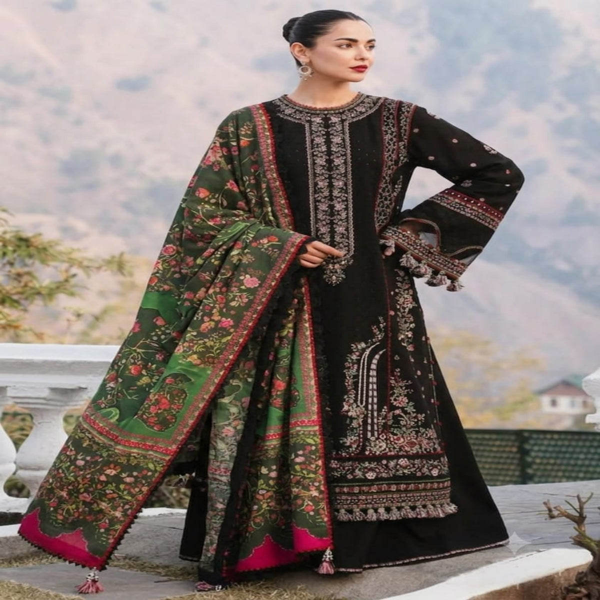 A woman models a traditional black and green embroidered ethnic suit with a matching printed dupatta, set against a blurred outdoor mountain background.
