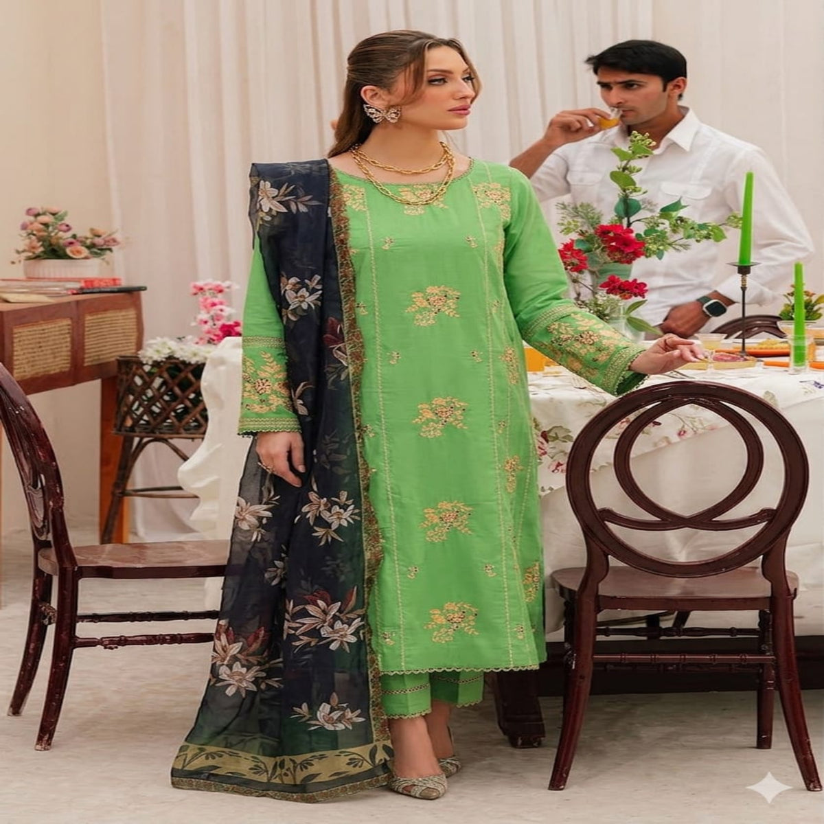 Woman in a green traditional outfit sitting at a dining table with a man in the background.