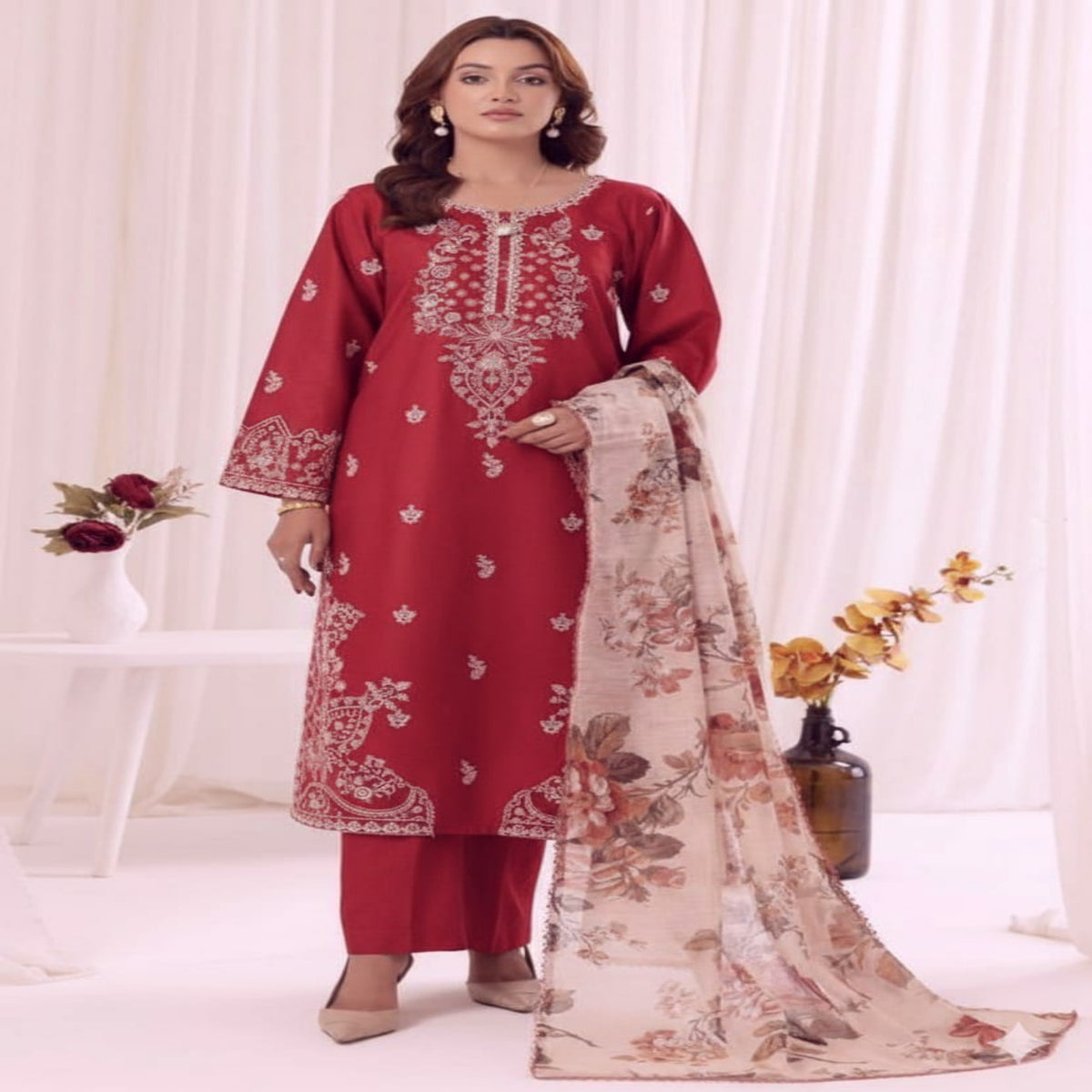 A woman models a traditional Indian salwar kameez suit in deep red with intricate silver embroidery, paired with matching red trousers and a beige floral dupatta, set against a soft white curtain backdrop.