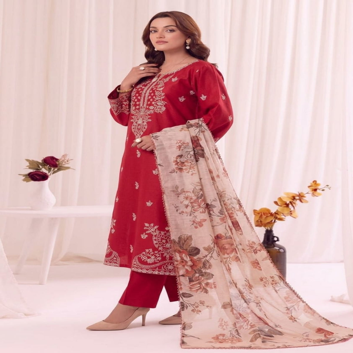 A woman models a three-piece red ethnic salwar kameez suit with floral embroidery and a beige floral dupatta, set against a soft white curtain backdrop with decorative floral arrangements.