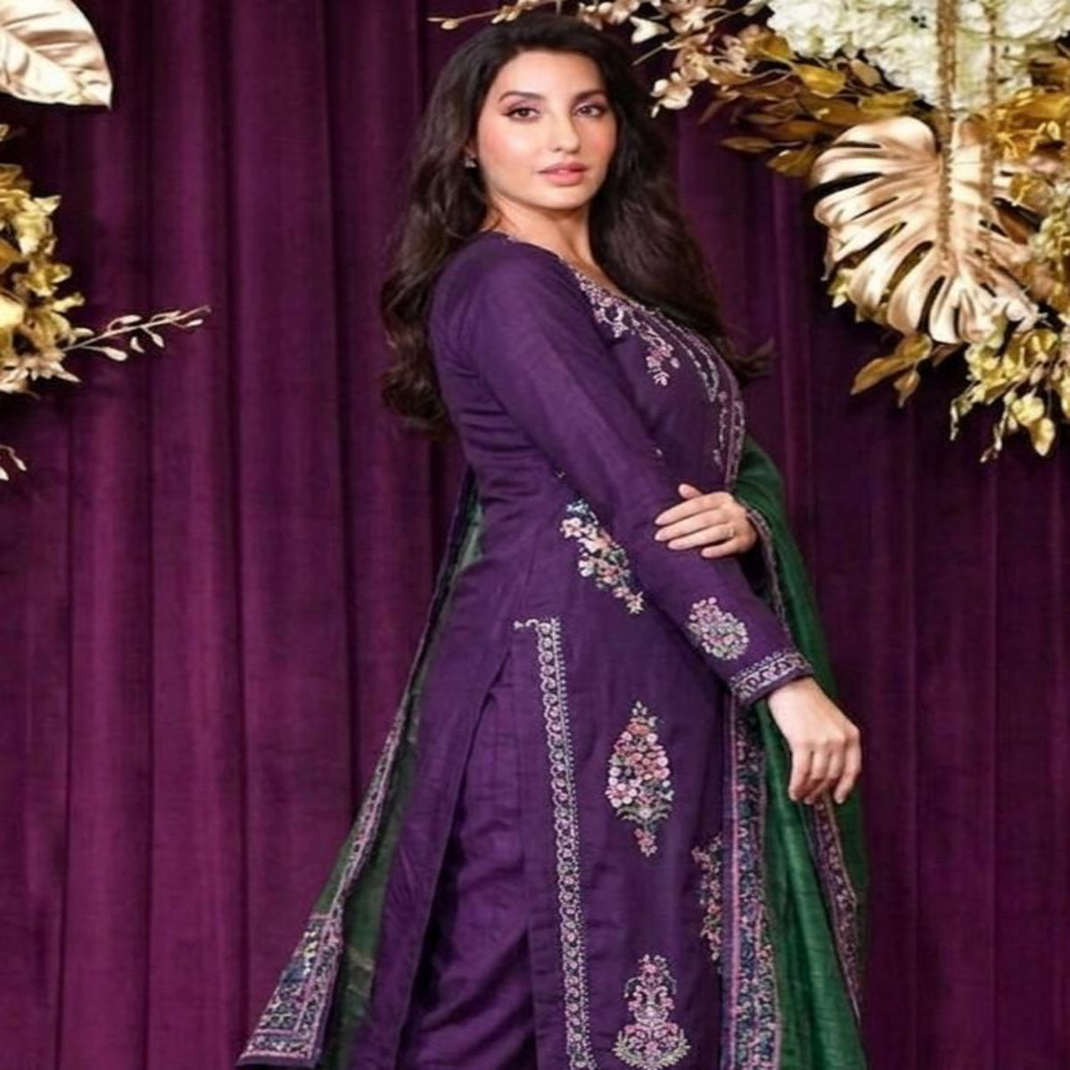 A woman modeling a traditional Indian outfit consisting of a deep purple kurta with intricate silver embroidery, paired with a dark green dupatta, set against a dark purple velvet curtain backdrop with gold decorative foliage.