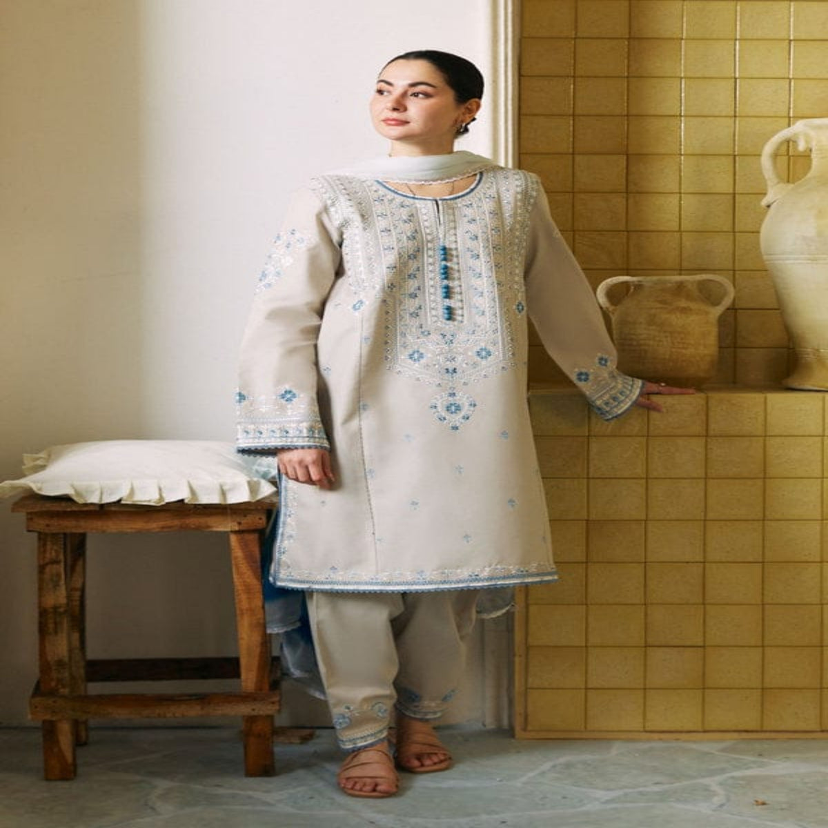 Woman in traditional outfit standing in a room with tiled walls and wooden stool.