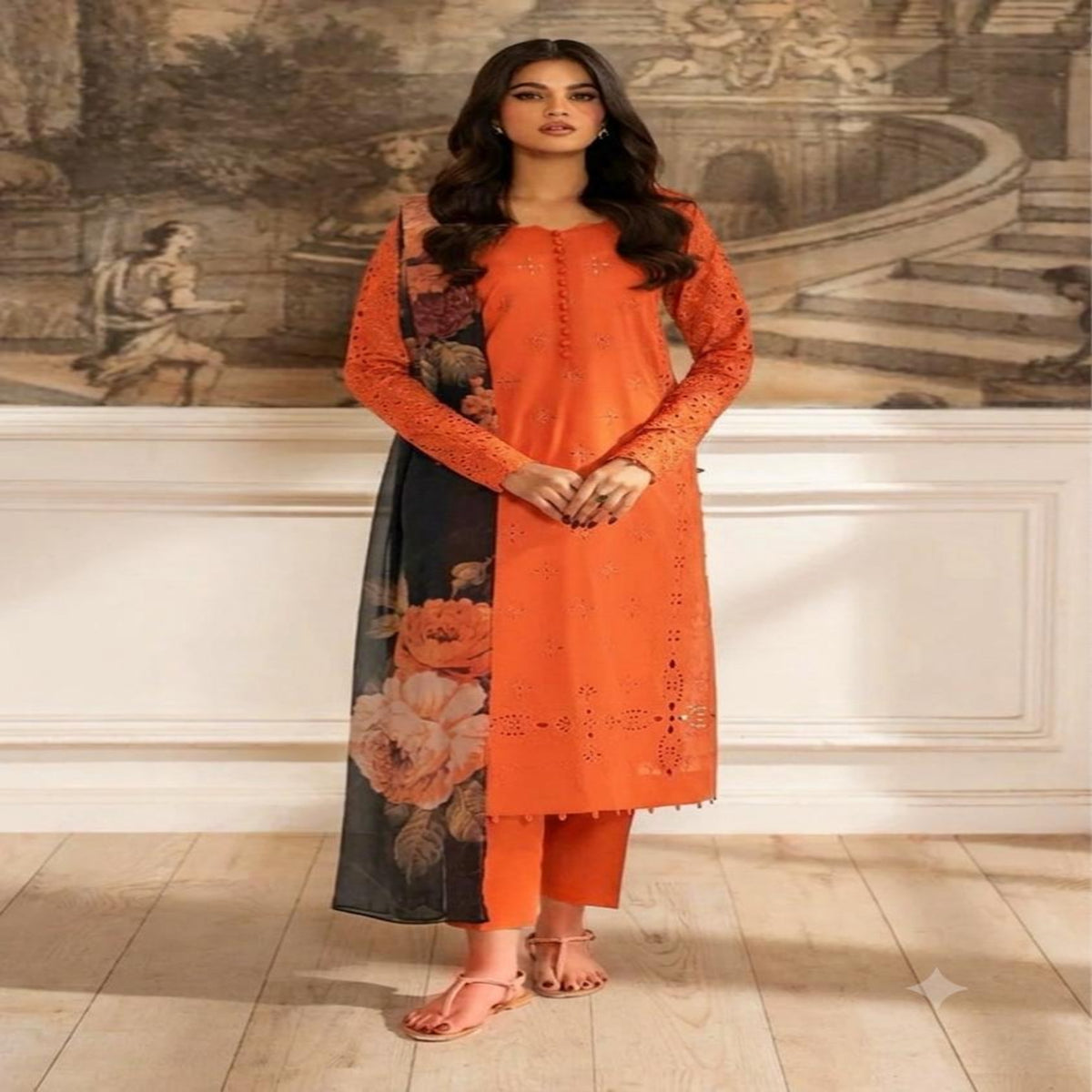 A woman models a vibrant orange kurta set with floral print panels, standing in front of a classical wall mural and light wood flooring.