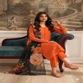 A woman models an orange salwar kameez suit with a black floral dupatta while sitting on a teal velvet armchair in front of a classical wall mural.