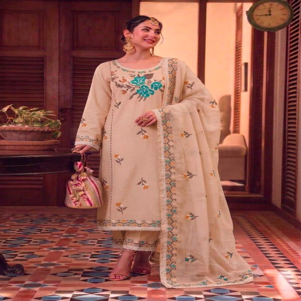 A woman models a traditional beige embroidered kurta set with a matching dupatta and pants in an indoor setting with patterned tile flooring and wooden doors.