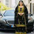A woman stands in front of a black car wearing a black traditional outfit with ornate gold embroidery and a matching embroidered dupatta. The background is a softly blurred urban street scene.