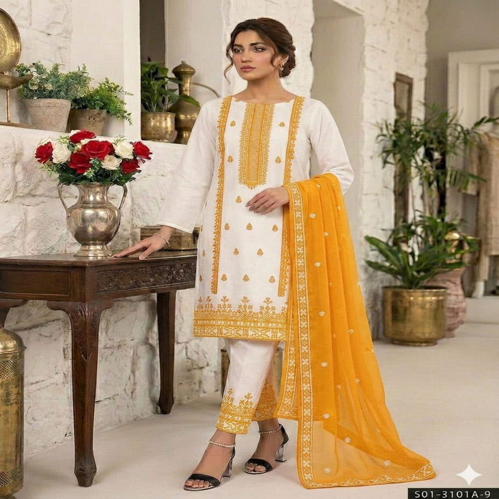 A woman models a traditional white salwar kameez suit with gold embroidery and a bright yellow dupatta in an indoor styled setting with plants and a wooden table.