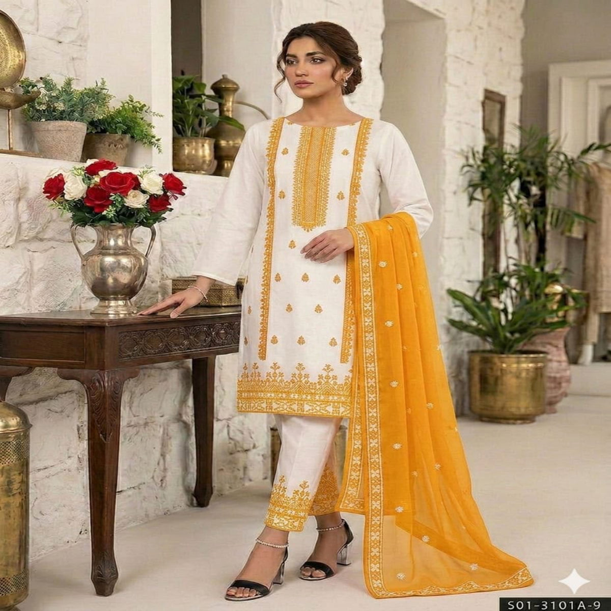 A woman models a traditional white salwar kameez suit with gold embroidery and a bright yellow dupatta in an indoor styled setting with plants and a wooden table.