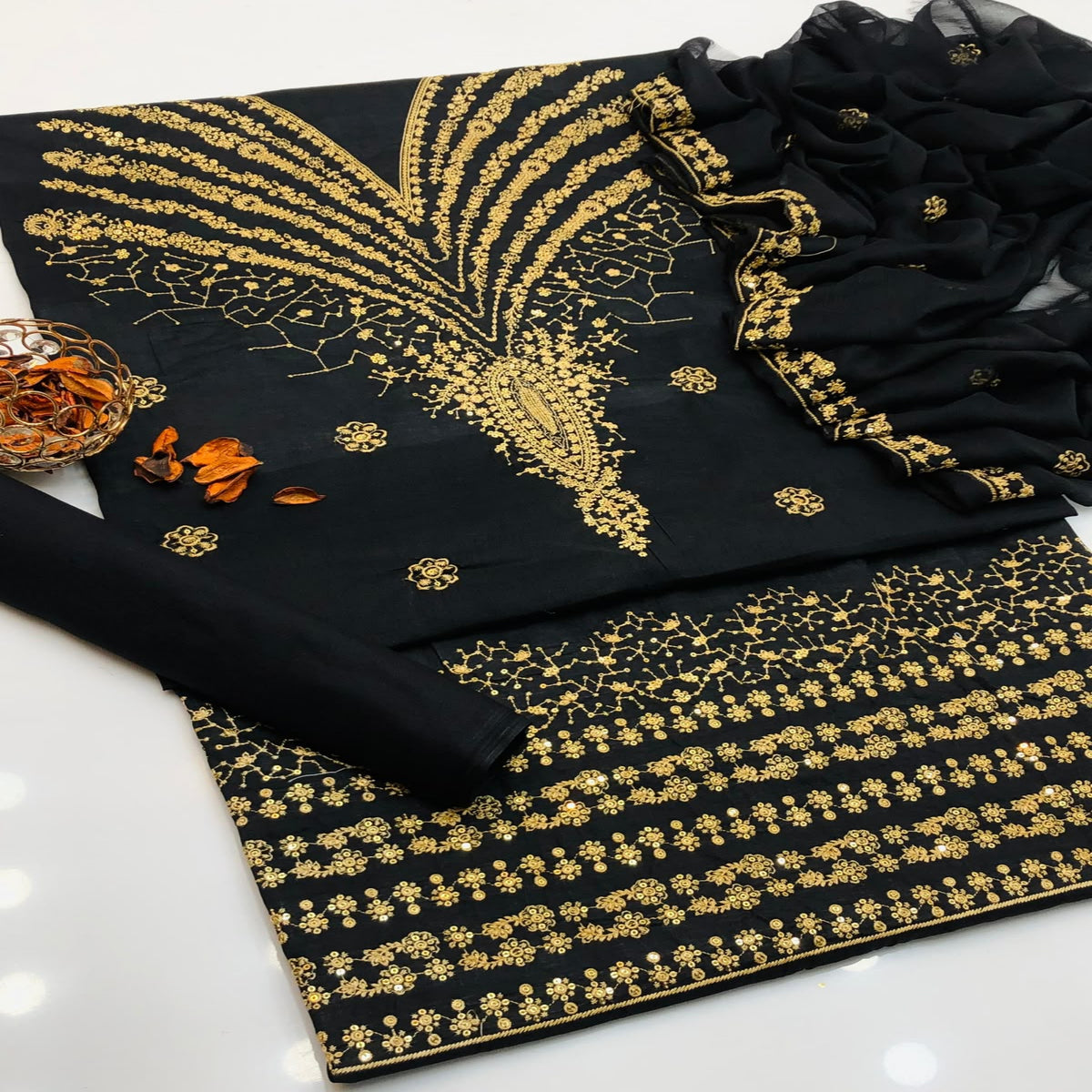 Black cotton fabric suit material with intricate gold zari embroidery, featuring a printed border, a matching dupatta, and a rolled fabric piece on a white surface.