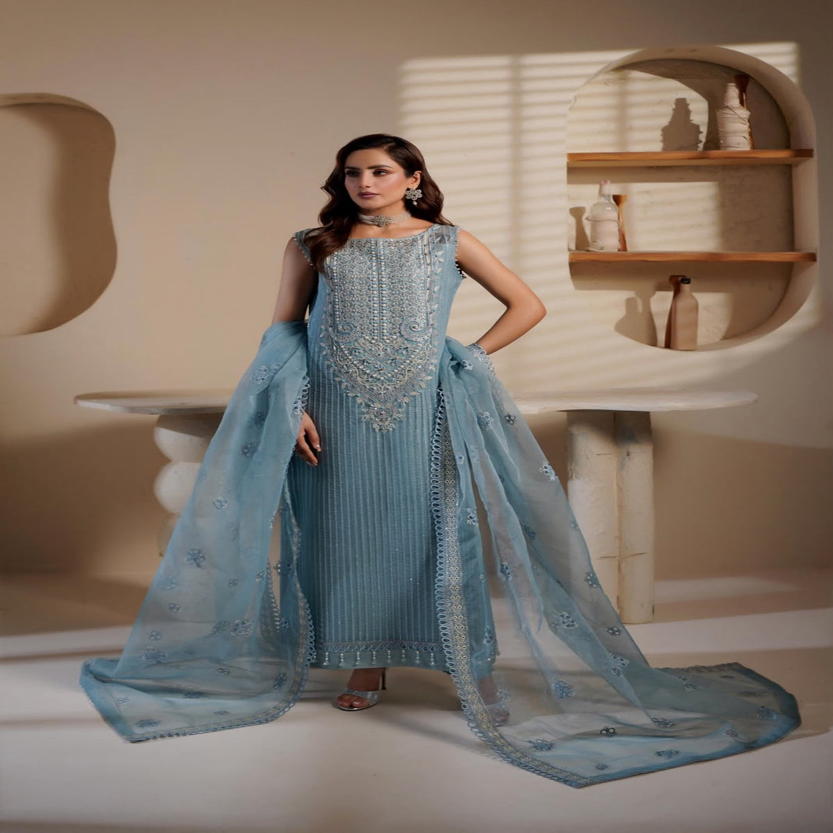 Woman in a light blue traditional outfit with intricate detailing in a minimalistic setting.