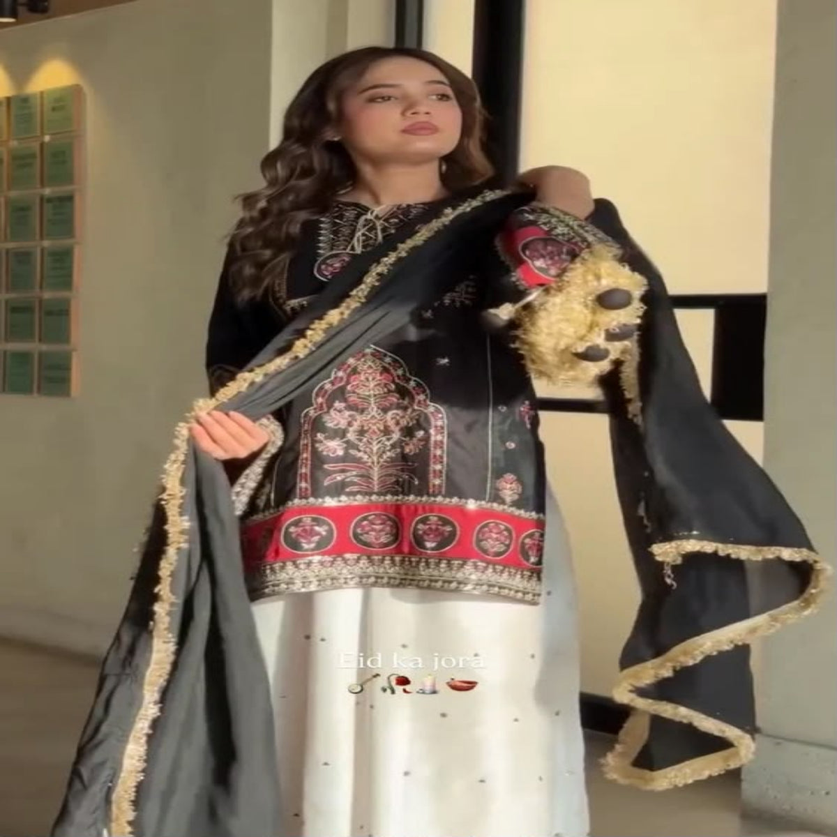 Woman wearing a traditional black and white outfit with intricate designs, standing indoors.