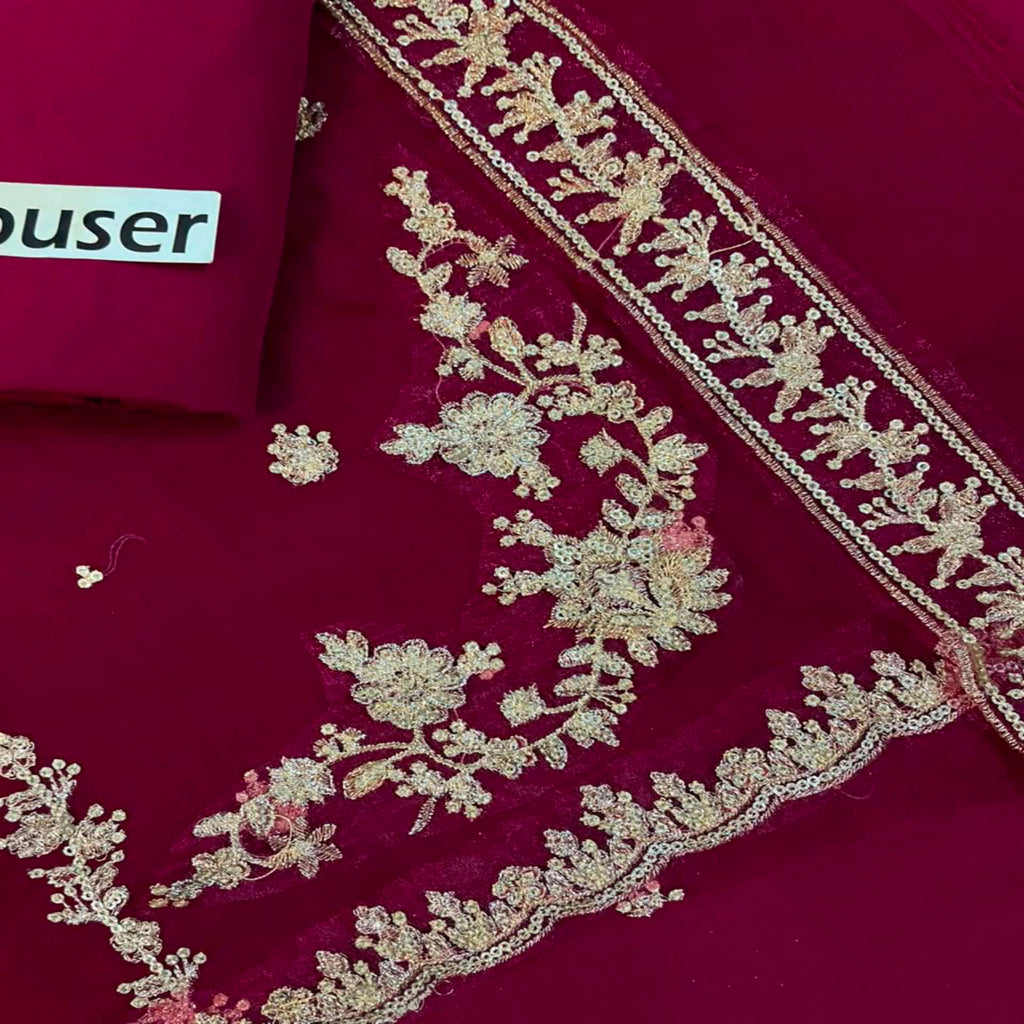 A close-up of a deep magenta or burgundy fabric with ornate gold floral embroidery and a matching embroidered border. A folded matching fabric piece in the upper left shows a partial white label reading 'user'.