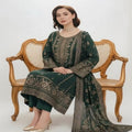 A woman seated on an ornate wooden cane-back bench wearing a dark green traditional outfit with extensive gold embroidery and a matching dupatta, photographed against a plain light background.
