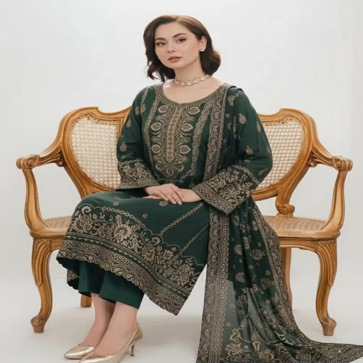 A woman seated on an ornate wooden cane-back bench wearing a dark green traditional outfit with extensive gold embroidery and a matching dupatta, photographed against a plain light background.