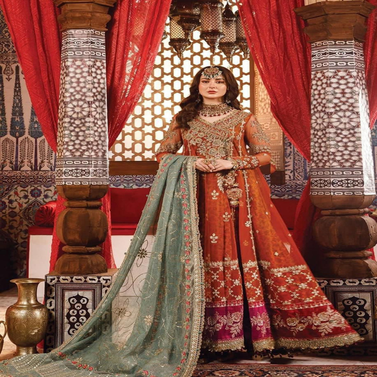 Woman in traditional orange and green embroidered outfit standing in a decorated room with red curtains and intricate patterns.