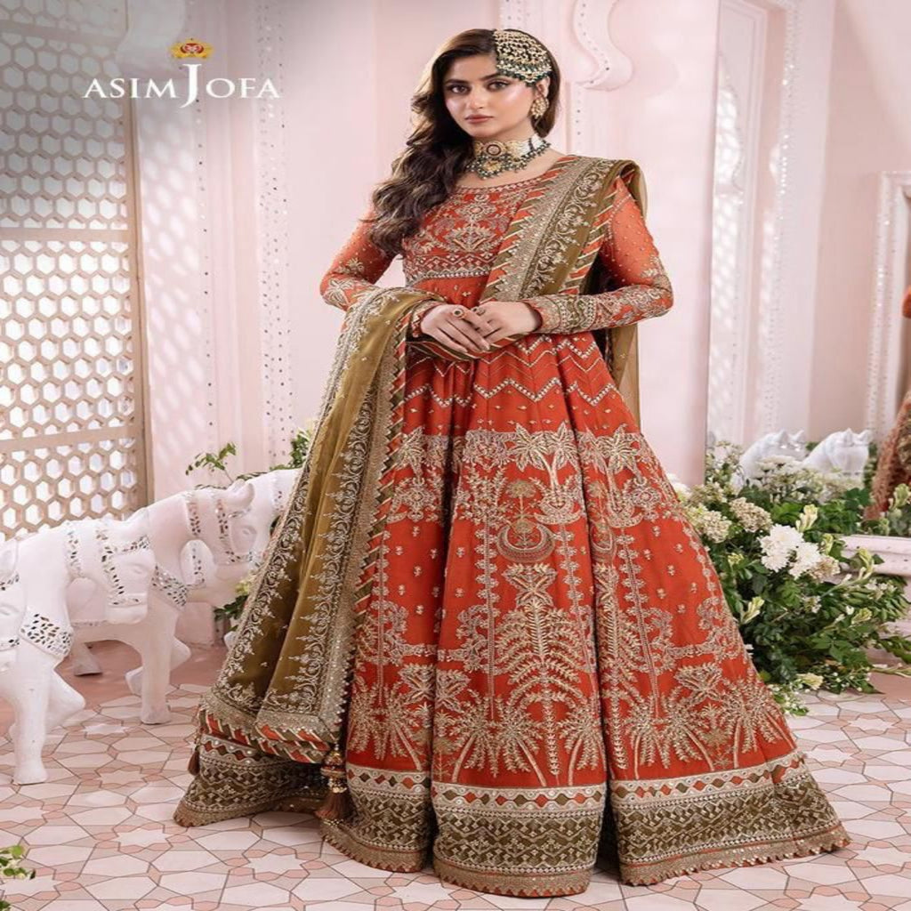 Woman in a traditional orange and green embroidered outfit with jewelry, standing in a decorated room.