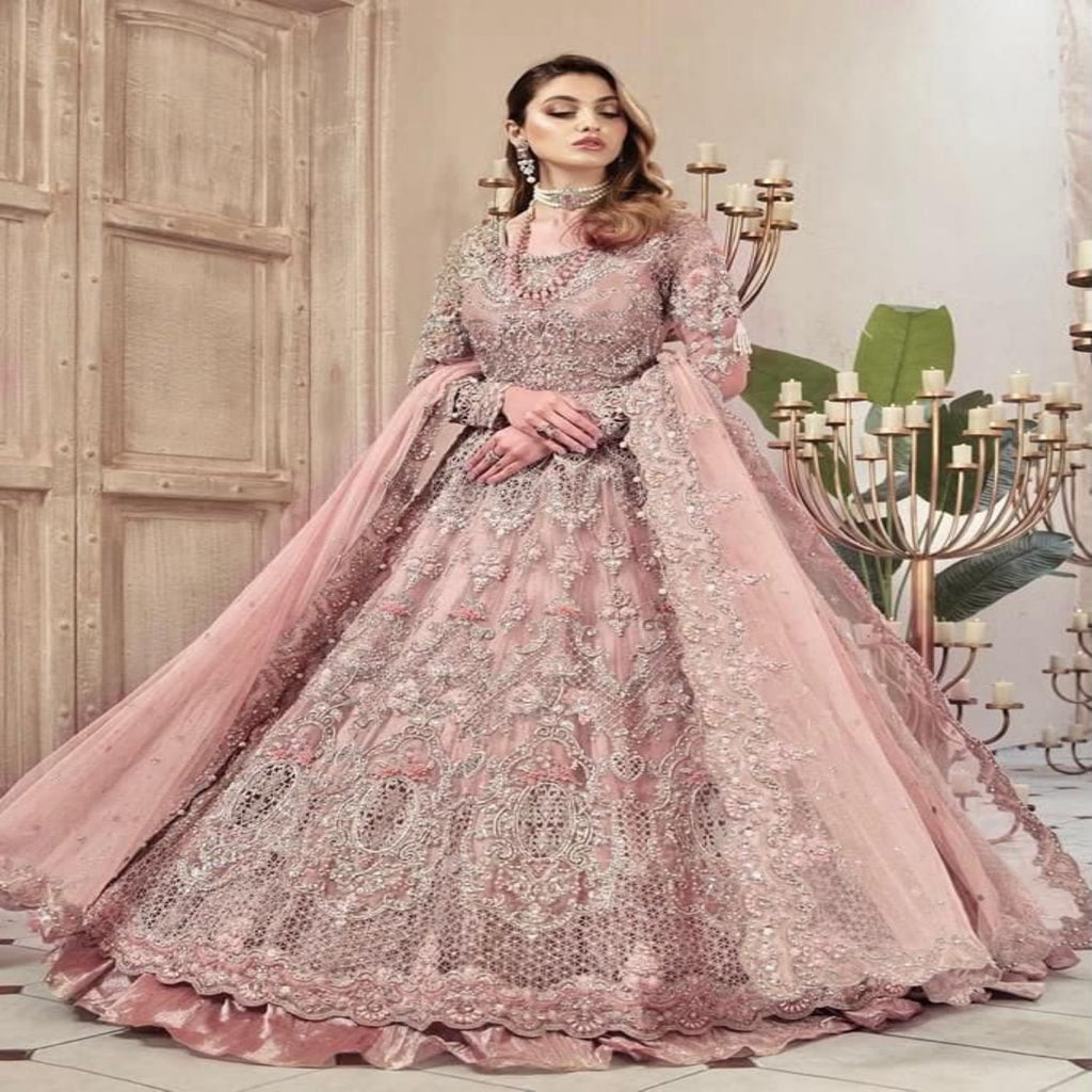 A woman models a voluminous pink embroidered traditional gown with a matching sheer dupatta, set against an elegant indoor backdrop with candelabras and a wooden door.