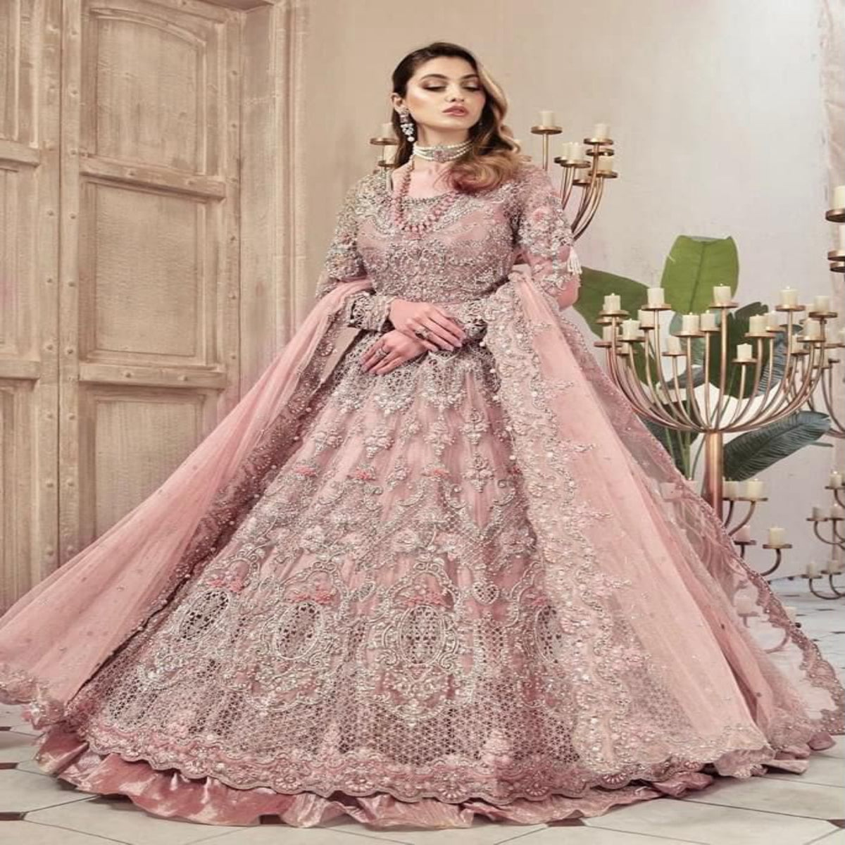 A woman models a voluminous pink embroidered traditional gown with a matching sheer dupatta, set against an elegant indoor backdrop with candelabras and a wooden door.