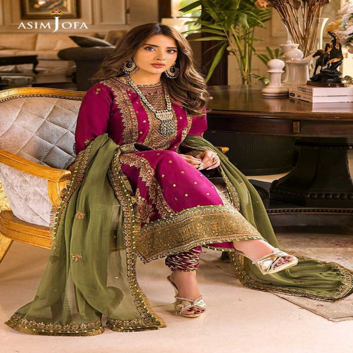 Woman in traditional pink and green embroidered outfit sitting in a luxurious room.
