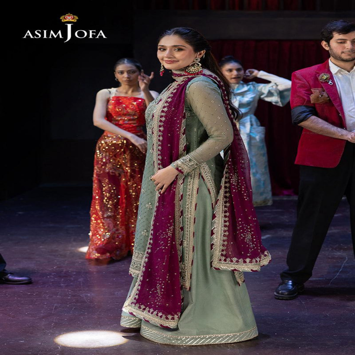 A woman modeling a traditional Indian outfit on a runway, wearing a sage green kurta with a deep magenta dupatta featuring gold embroidery. Other models and a man in a red blazer are visible in the background under stage lighting.