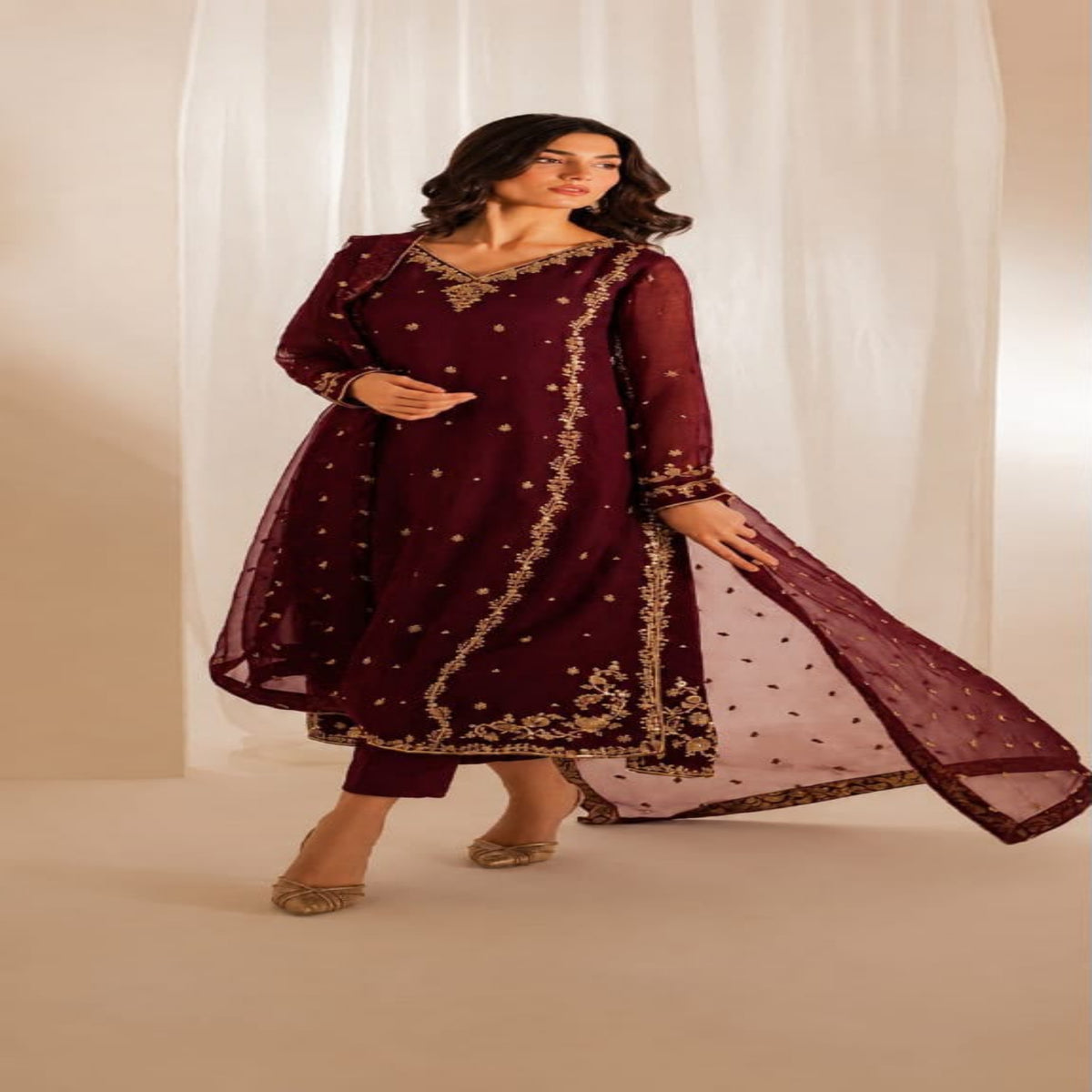 A woman models a traditional Indian outfit consisting of a deep maroon kurta with gold embroidery, matching maroon trousers, and a sheer maroon dupatta with gold polka dots, set against a warm beige background with white drapes.